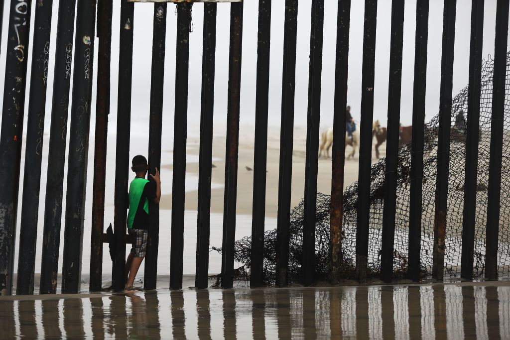 A boy stands between posts in the U.S.-Mexico border fence, with people riding horses visible on the American side, on a beach in Tijuana, Mexico, on March 9, 2018. (Mario Tama/Getty Images)