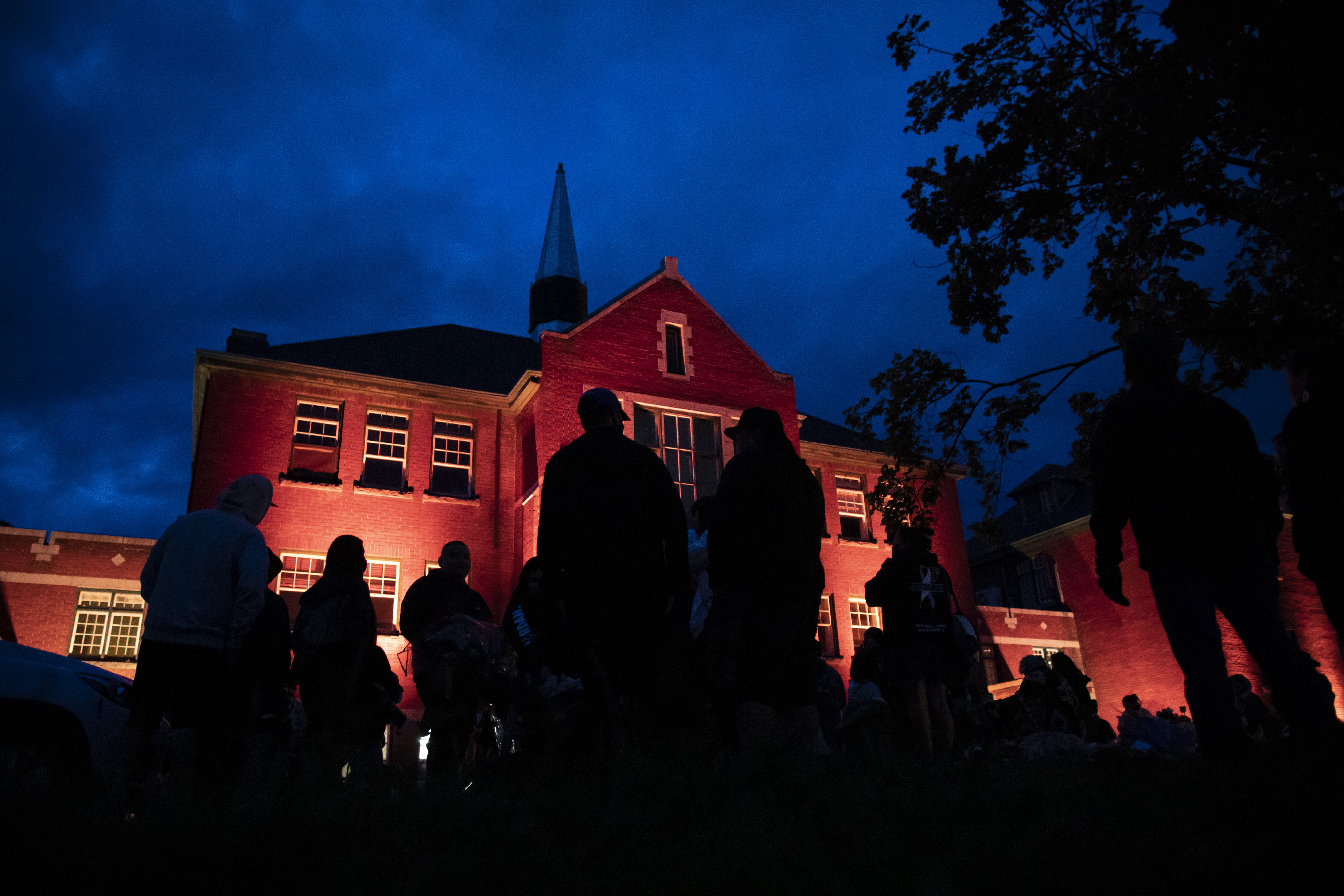 The former school is illuminated with orange lights in the evening as people gather outside the memorial.