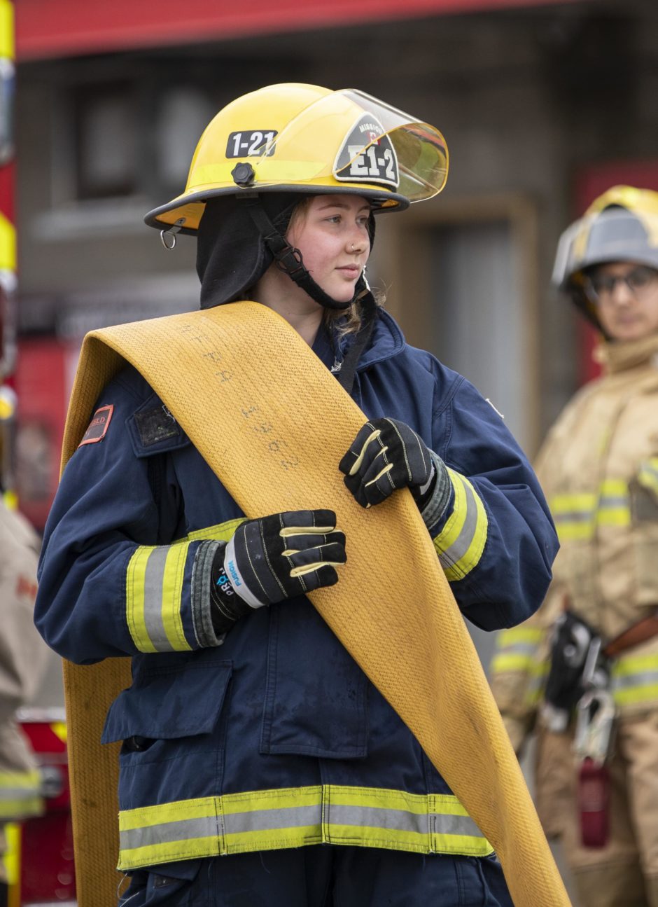 A teenage girl in firefighting gear holds a firehose. 