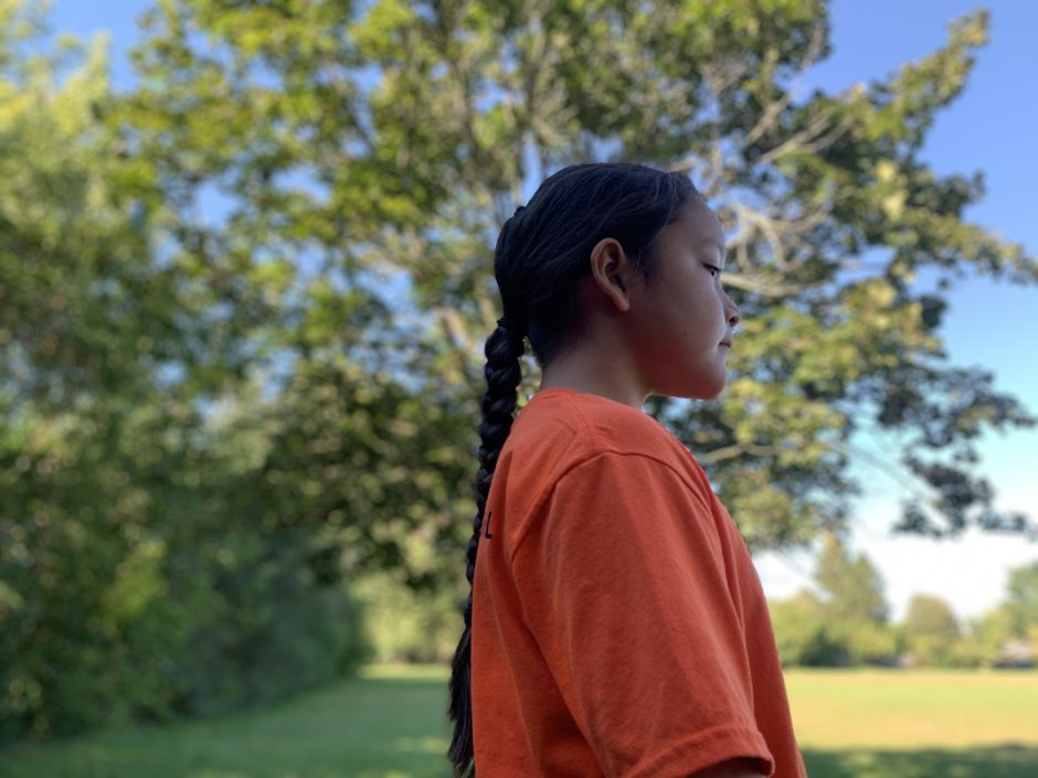 A nine-year-old Indigenous boy with a long braid stands staring into space wearing an orange t-shirt. (Kate Dubinski/CBC)