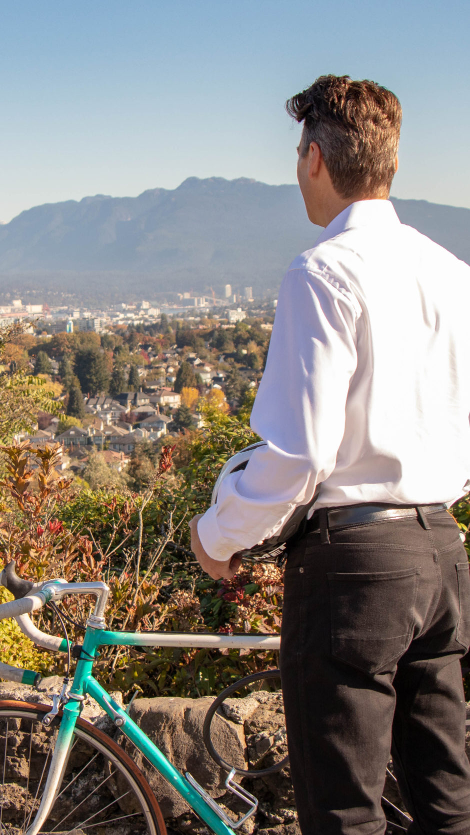 Gregor Robertson is looking at the skyline while standing next to a bicycle. 