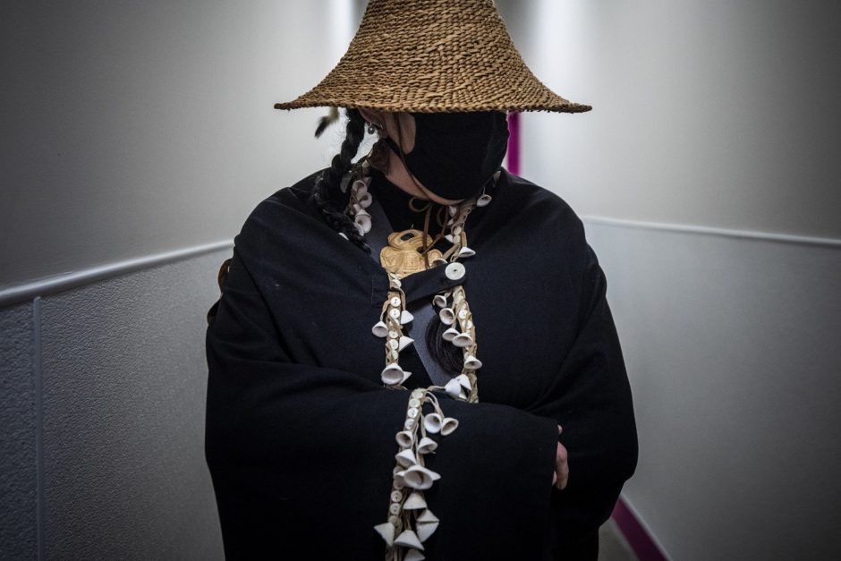 A woman stands draped in a traditional Haida shawl and woven basket hat with her face hidden behind a black mask.