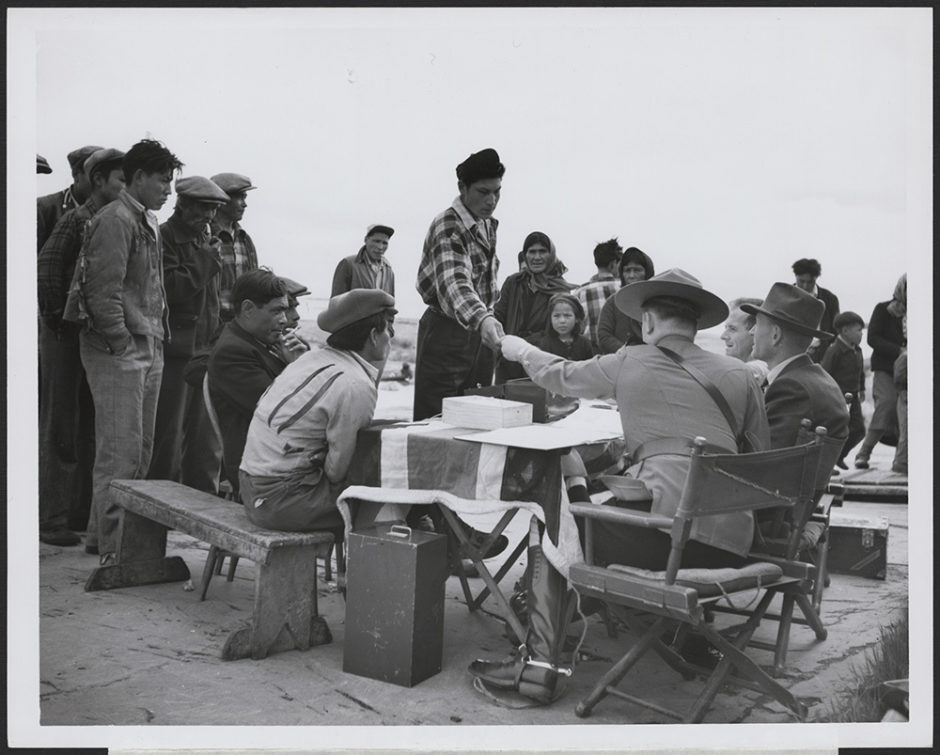 A Dene man accepts money from a fully uniformed Mountie seated behind a table draped with a British flag. 