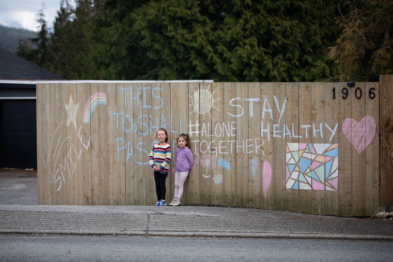 Presley Muir, 4, and Scotia Muir, 6, stand in front of a chalk sign they drew while trying to keep busy over the weekend in North Vancouver on Monday, March 23, 2020. (Maggie MacPherson/CBC)