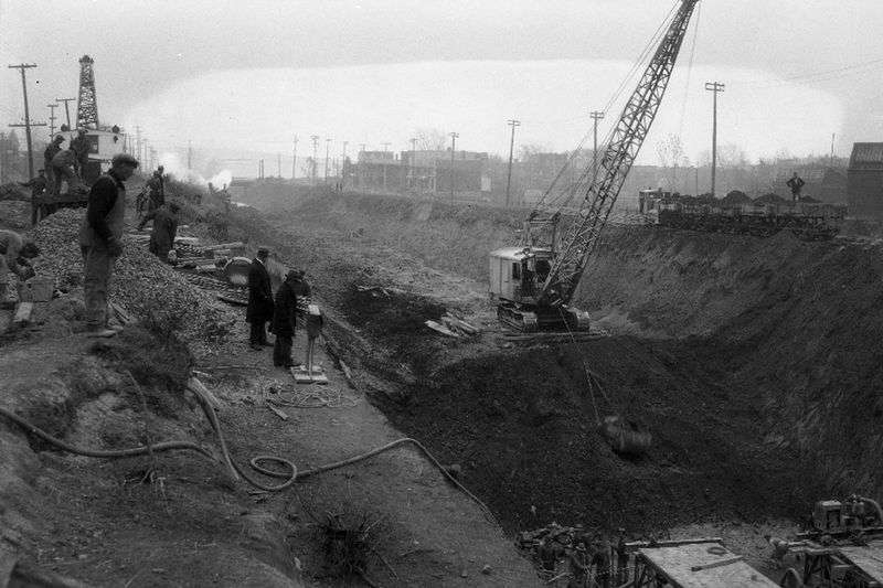 A black and white photo of a construction crane in the middle of a canal. There are workers and observers standing on the side of the canal.