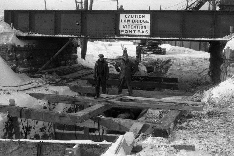 Two men stand on wooden beams balancing across a channel. There is snow on the ground. A bridge with a sign reads: Caution low bridge. Attention pont bas.