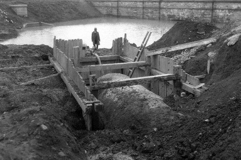 A black and white photo of a partially buried concrete culvert surrounded by wood framing. There is standing water in the background and a man.