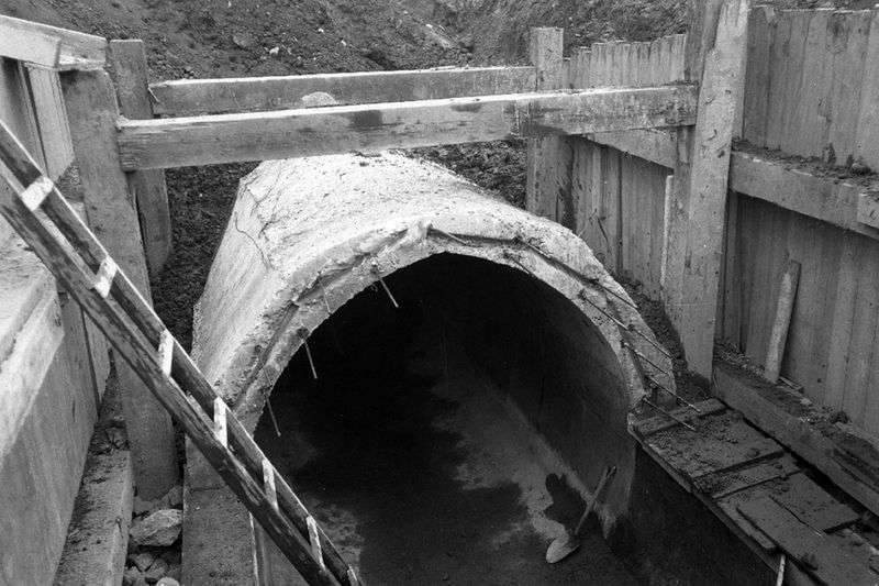 A black and white photo shows an empty concrete sewer pipe, partially buried under dirt. A wooden ladder leads out of the dug channel.