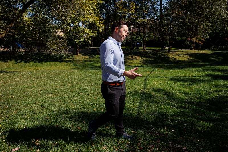 A man with short brown hair and a blue button-up shirt walks through a park. His hand is gesturing.