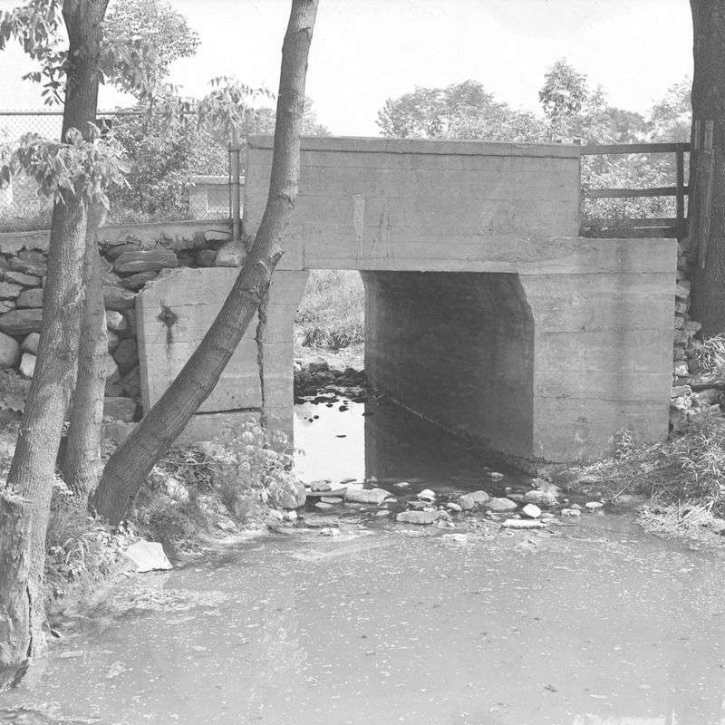 A black and white photo of a stone and concrete bridge over a shallow creek.