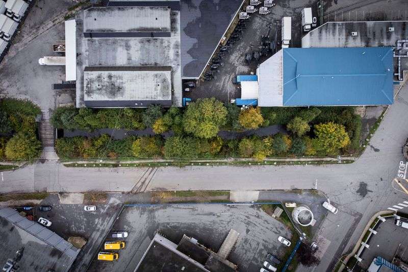 A creek seen from above runs parallel to a road, through a green strip of land bordered by industrial buildings and pavement.