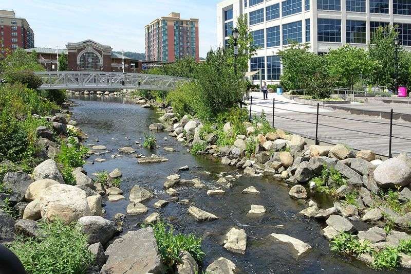 A waterway flows through a city. Its banks are bordered by rocks and vegetation. There is a bridge over the water.