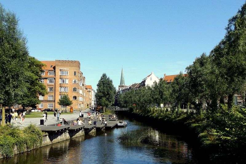 People sit on raised platforms, overlooking a river that flows through a city. There are homes and a church steeple in the background.