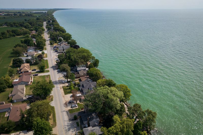 An aerial view shows a road running parallel to a lake shore. The home is lined by homes. On one side of the road, the homes back onto the water. The water is greenish blue.