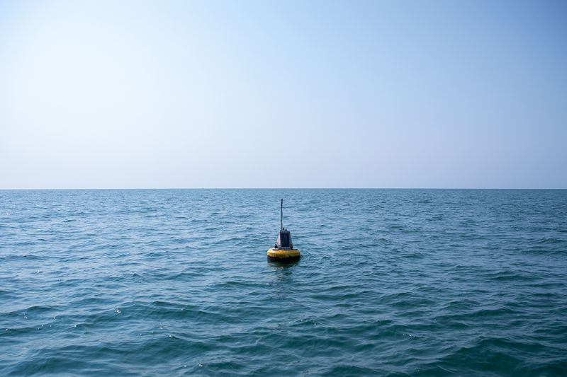 A yellow buoy floats on the surface, surrounded by an expanse of water and sky. A grey and black rectangular object and an antenna are mounted on top of the buoy