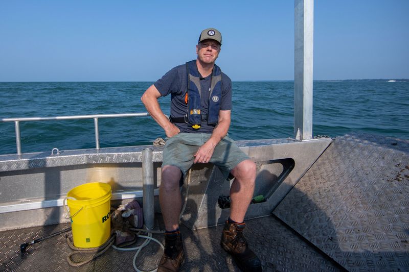 A man wearing a ballcap, shorts and a t-shirt sits on the edge of a metal boat, looking towards the camera. He’s seated casually, with one arm on his hip. There’s a bucket and rope on the floor next to him. The background is filled with a large swath of blue water and blue sky.