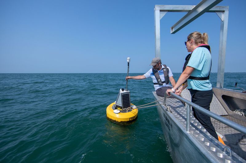 A man leans over the edge of a boat to grab a buoy floating on the water. A woman standing next to him is holding a rope that is attached to the buoy, helping to reel it in.