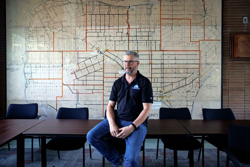 A man sits on a board room table. His gaze is off to the side of the camera. He has a beard and short hair that’s greying. He wears glasses, a black short-sleeved polo shirt with a Union Water Supply logo on the breast, and blue jeans. Behind him, a large map of the region covers the wall. The map, which seems to be vintage, is divided into grids with labels like Tilbury North, Tilbury South, and Kent.