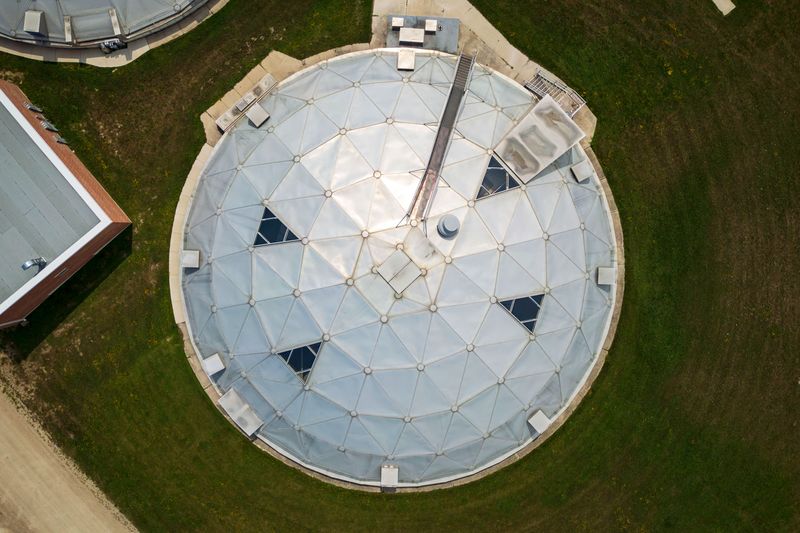 A top-down view of a large silver-coloured domed building, seen from outside. There are four triangle shaped skylights in the building’s curved roof and stairs that lead up to the side of the building. The grass around the building is mowed.