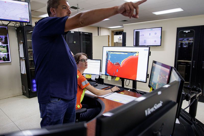 A man in a blue short-sleeved polo shirt stands and points to something off-camera. He is inside a room with fluorescent lighting. There are monitors mounted on every wall and on the desk in front of him. A woman sits at a desk next to him and looks in the direction the man is pointing. She’s wearing an orange Hi-Vis safety shirt and has her hand on a computer mouse. The screen in front of her is displaying a website that shows the outline of Lake Erie.