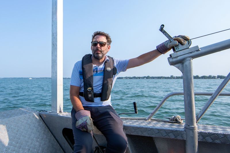 A man sits on the edge of a boat, wearing sunglasses, a polo shirt and slacks. His gaze is directed off camera. He’s wearing thick work gloves and one of his hands is resting on a winch. In the background, the lake extends far into the distance, meeting the shoreline and a blue sky.