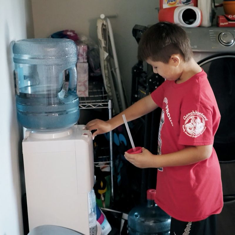 A boy presses the button on a water cooler to pour water. The water cooler is in a room next to a laundry machine with other household storage.