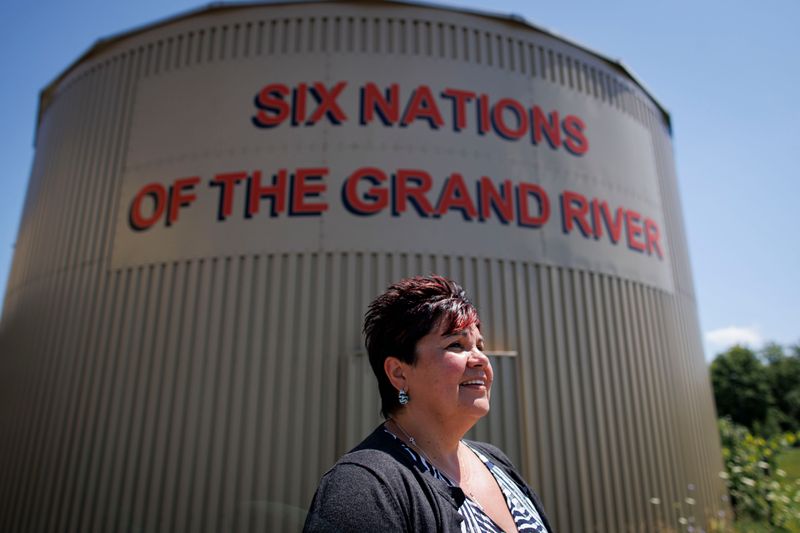 A smiling woman with short red hair stands outside. The sun lights her face. Directly behind her there is a cylindrical tower. A sign on the structure reads “Six Nations of the Grand River.”