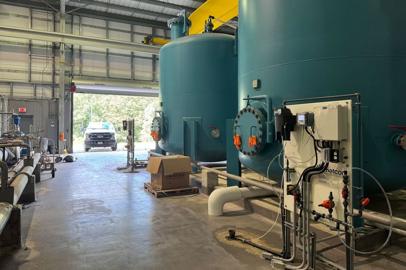 A view inside a water treatment plant shows the interior of a large building with high ceilings, filled with tanks connected to pipes and conduits. The floor is cement slab. A garage door is open, revealing a parked pickup truck.