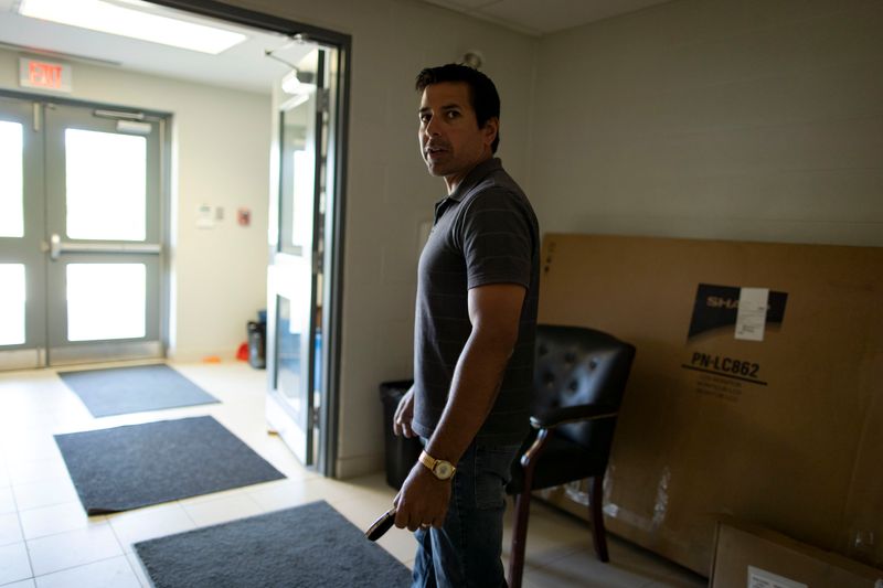 In a wide hallway with fluorescent lighting and tile flooring, a man looks back over his shoulder mid-stride. In front of him sunlight streams through the building’s entrance doors.