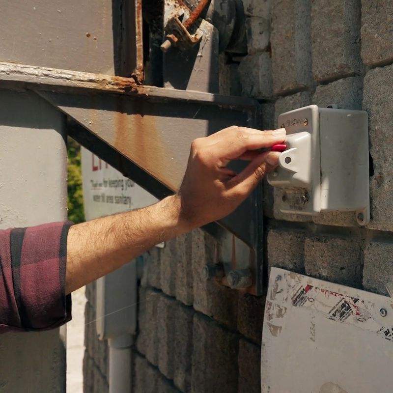 A closeup image shows a man’s hand flicking a switch attached to the outside of a concrete wall. Pipes and metal fixtures — some of them rusted — jut out of the wall in the background.