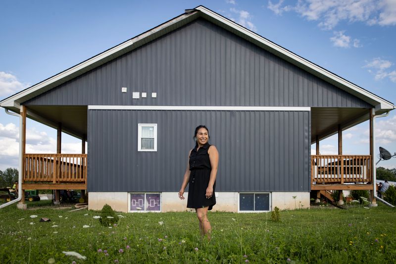 A woman stands outside on the grass under a blue sky. The lawn is blooming with clover and wildflowers. Behind her is a modern dark blue bungalow, with a front and back porch. Inside one of the basement windows is the Iroquois confederacy flag, which is purple with four connected white squares and an eastern white pine tree in the centre.