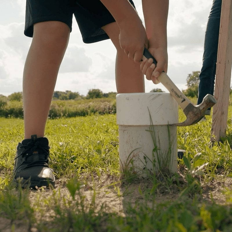 A closeup view shows a grey tube sticking out of the grass. The claw end of a hammer, held by a boy’s arms, is being used to pry a white cap off the tube.