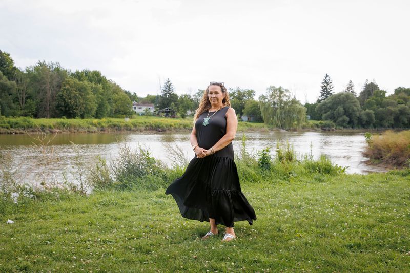 A woman stands posed outside in short grass, with her hands clasped in front of her. She’s wearing a long black summer dress and her expression is solemn as she looks into the distance. Behind her there is tall grass lining the shore of a river. In the background there are some homes on the other shore.