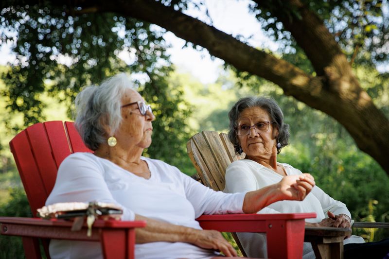 Two senior women sit outdoors in the shade in muskoka chairs, placed side by side. They’re surrounded by greenery and trees. One woman appears to be in the middle of speaking and the other woman’s head is turned to look at her.