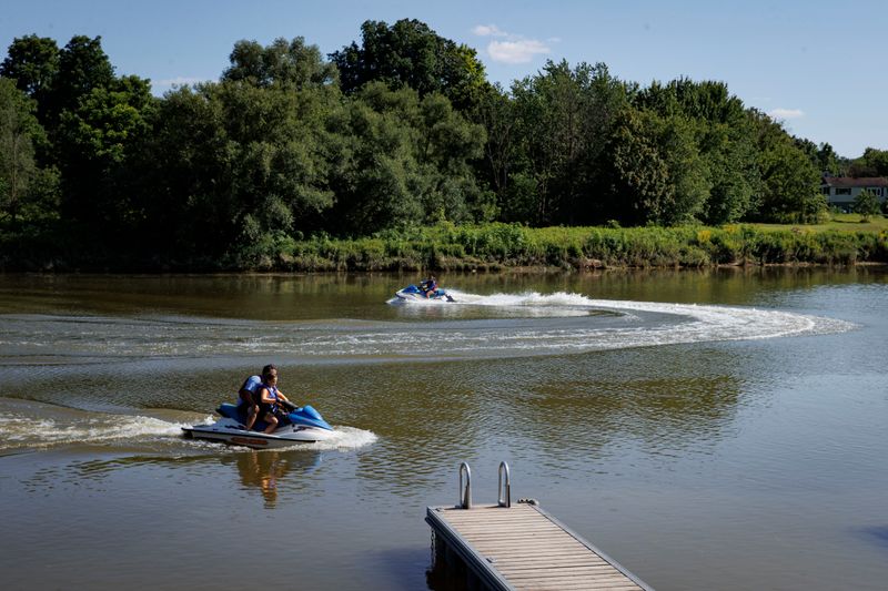 Two jet skis circle a murky river, leaving a wake behind them. A wooden dock juts out into the water. The shore is lined with tall grass and trees.