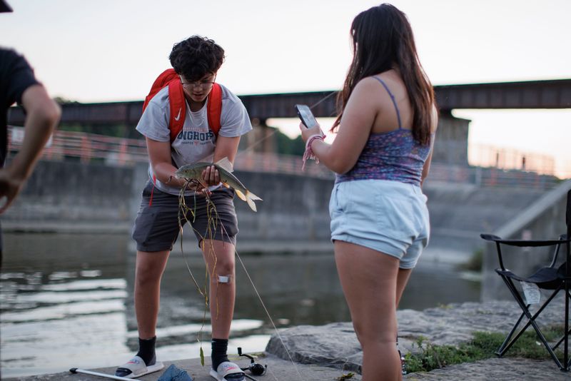 A young adult holds a fish in their hands on the shore of a river as a friend snaps a photo with their cell phone. The shoreline is lined with concrete walls and stone.