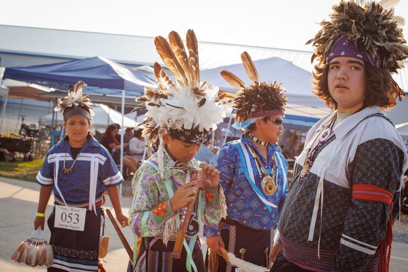 Four boys stand in a group outside, adjusting their traditional dress. They’re wearing feathered headpieces and clothing with beaded and woven embellishments. In the background, pop up tents shade a crowd of people sitting on folding chairs. One of the boys has a large label pinned to his shirt that said “Grand River Smoke Dance.”