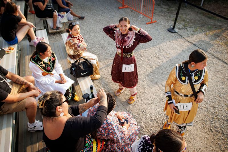 A group of five women wearing traditional dress, some sitting and some standing, are gathered in a crowd around bleachers outside. They’re wearing moccasins and brightly coloured clothing that’s embellished with embroidery and ribbons. Two of the women have labels pinned to their clothing that say “Grand River Smoke Dance.”