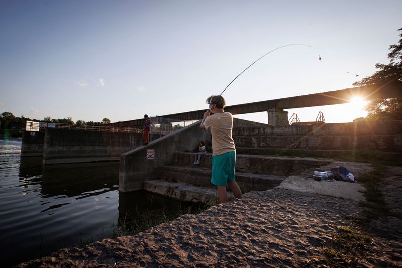 A young adult wearing headphones casts a fishing line into a river as the sun sets. The shore they are standing on is concrete. There’s an old defunct dam in the background and a sign that warns: Danger, No Swimming.