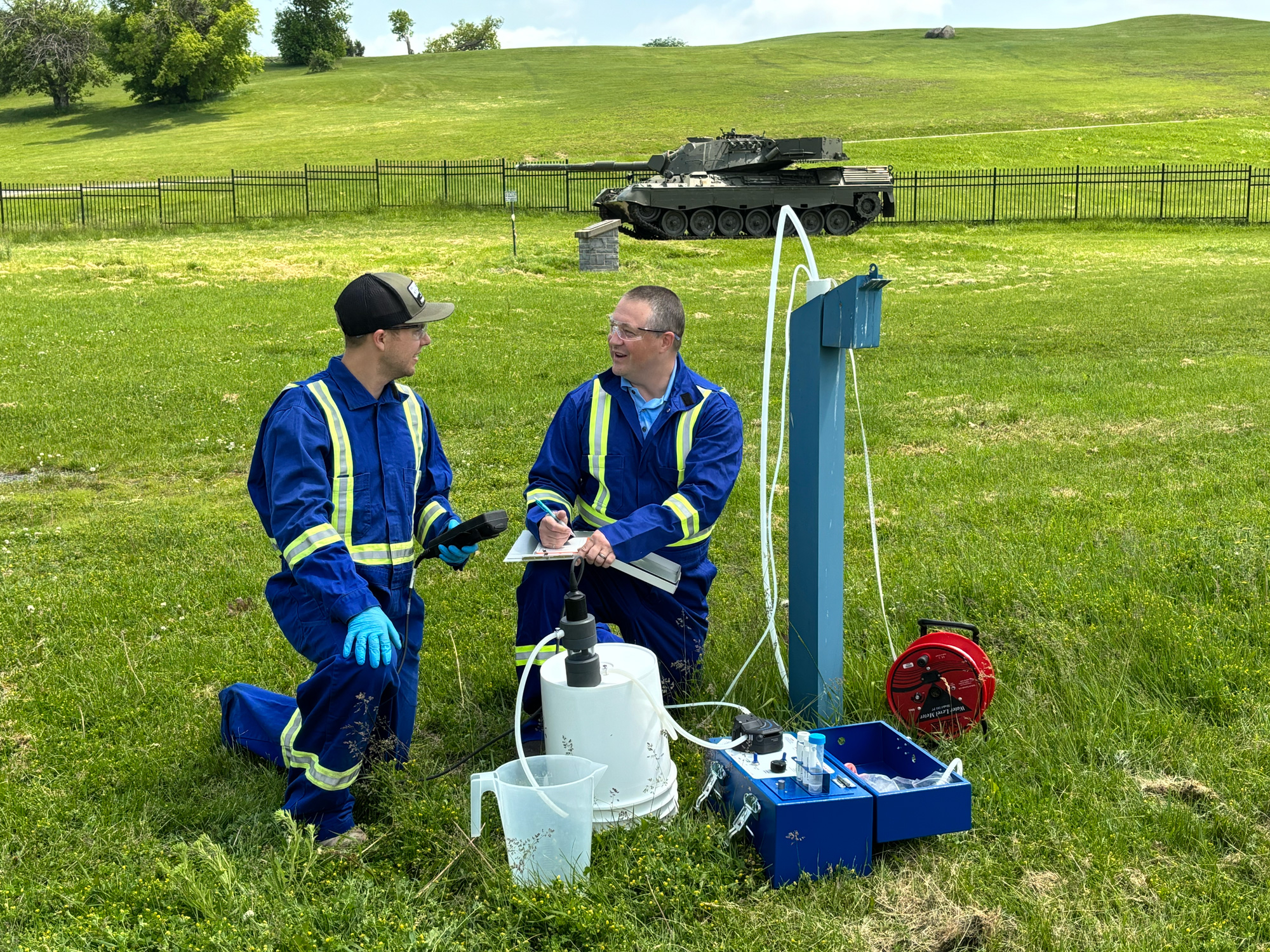 Two men kneel in a grassy field, both wearing safety glasses and blue coveralls adorned with reflective tape. The man on the left is wearing a black baseball cap and holding a black device in his hand. The man on the right has a buzzcut and is writing on a notepad that is balanced on his knee. There are various sorts of equipment next to them, including tubing coming out of the ground and what appears to be a hand pump, a blue toolbox and test tubes. There is a military tank in the background.