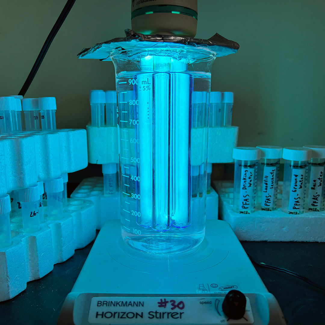 A man with a buzzcut wearing safety goggles looks through the glass window of a hood vent in a lab. He is holding a test tube in his left hand that says 'PFAS – groundwater.' Inside the hood vent there are stacks of test tubes and in the middle, a cluster of bright blue vertical lights have been submerged in a glass pitcher of water. On the front of the hood vent, a sign states, 'Reminder: All containers must be labelled with contents, date and name. Containers being used temporarily or as transfer containers must also be labelled.'