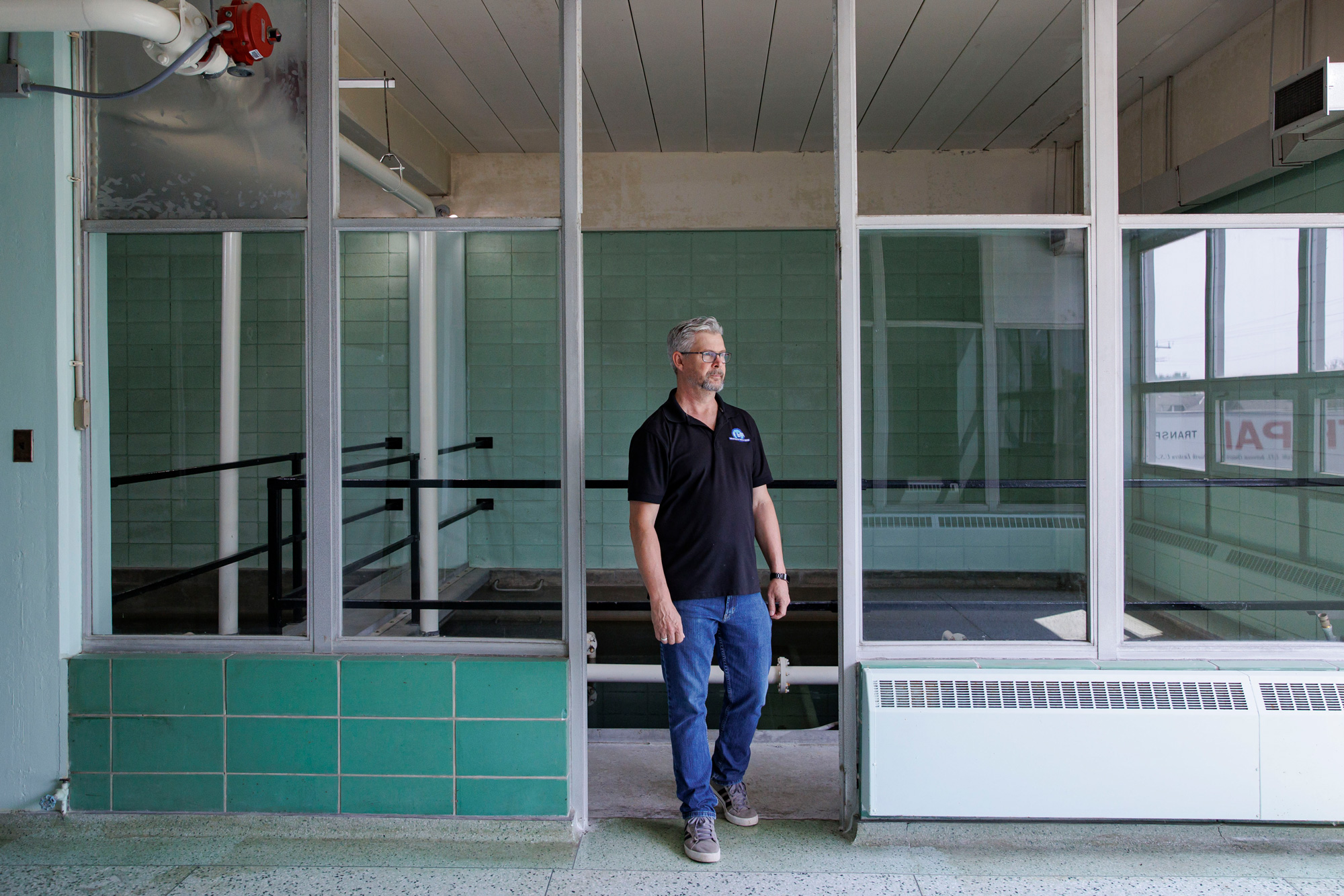 A man wearing a black polo shirt, jeans and sneakers walks through an interior doorway. He has short grey hair and a trimmed beard. He is wearing glasses. There is green tile on the wall behind him and a staircase leading down. Either side of the doorway is panelled glass.
