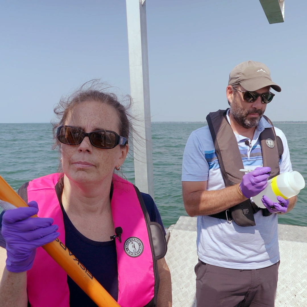 A photo shows two people standing side by side on a boat, surrounded by water. On the left, a woman wearing sunglasses and a pink life vest looks toward the camera and holds an orange pole. To the right, a man wearing a baseball cap, sunglasses and a grey life vest looks down at a canister he’s holding. The canister is filled with water and has a yellow label, which he is writing on with a Sharpie marker.